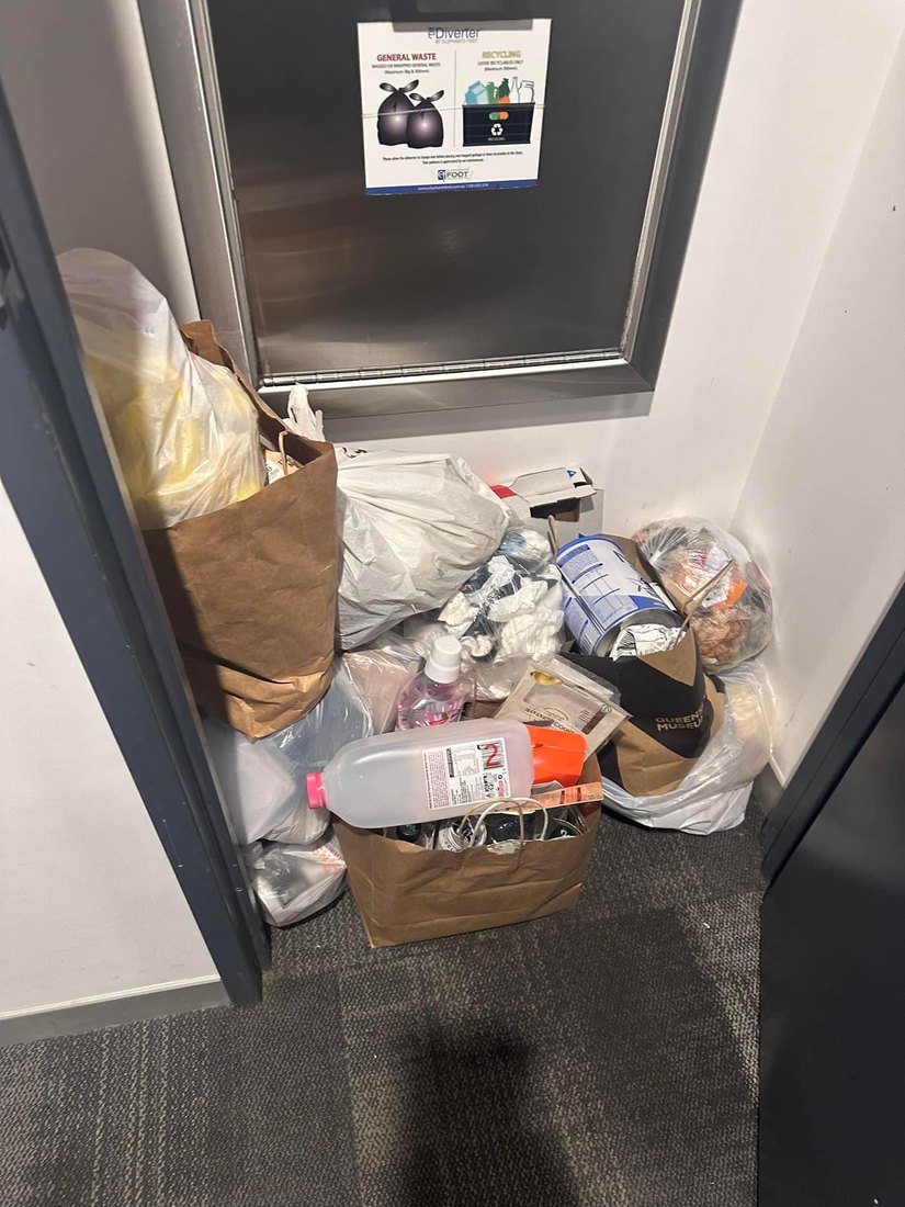 Bags of food waste and rubbish piled beside a lift in a Brisbane apartment building, next to a recycling and general waste sign.